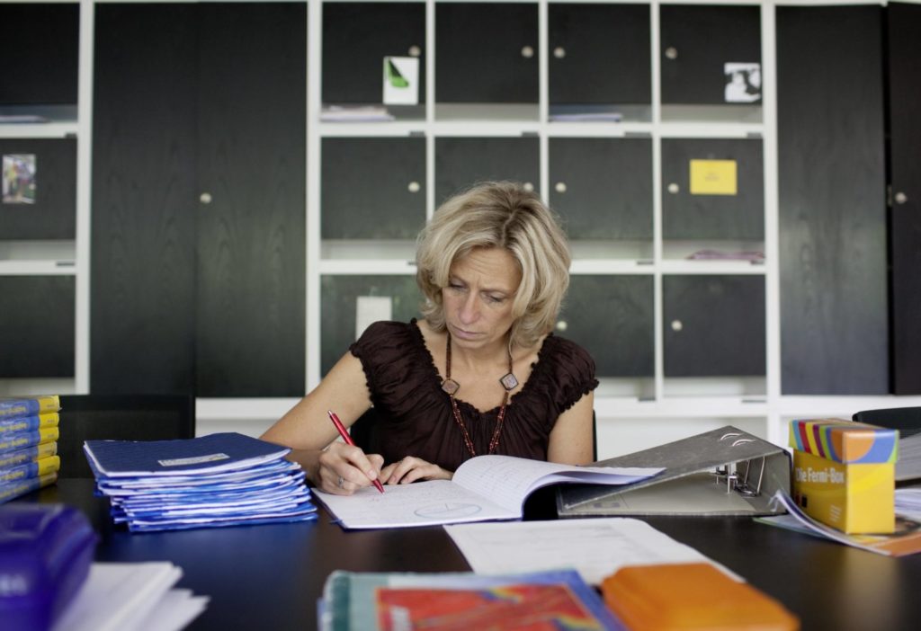 Man sieht eine Frau die an einem Schreibtisch voller Ordner und Hefter sitzt und schreibt: Verschlechtern Quereinsteiger die Qualität des Unterrichts an Schulen?