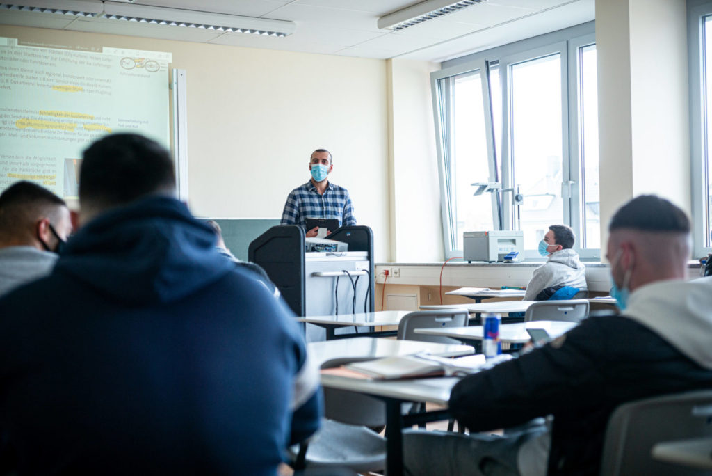 Bild von einem Klassenzimmer, man sieht Menschen an Tischen sitzen und eine Person am Sprecherpult: Bisher gibt es kaum Sprachförderung für Berufsschullehrer