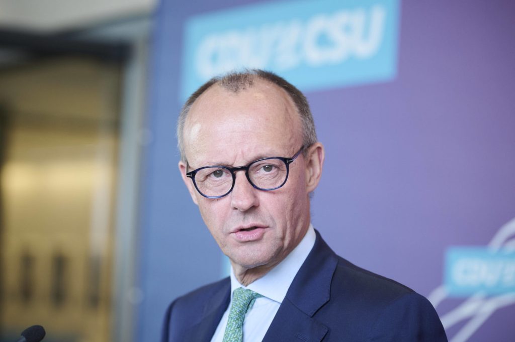 Porträtfoto von Friedrich Merz mit blauem Anzug und Brille vor einer blauen Plakatwand der CDU/ CSU