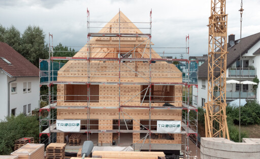 Ein Haus aus Holz wird gebaut, daneben ein Kran, im Hintergrund weitere Häuser