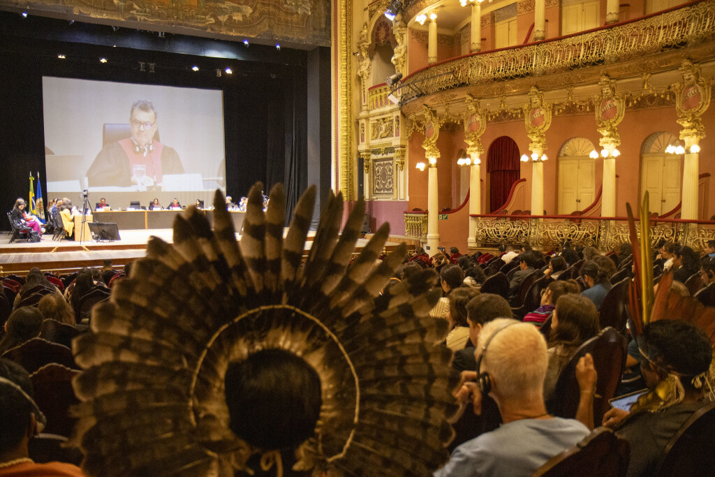 Blick aus dem Zuschauerraum auf die Bühne eines prunkvollen Theaters. Auf ihr tagt ein Gericht. Im Vordergrund sieht man den Kopfschmuck einer indigenen Person, die an der Anhörung teilgenommen hat.