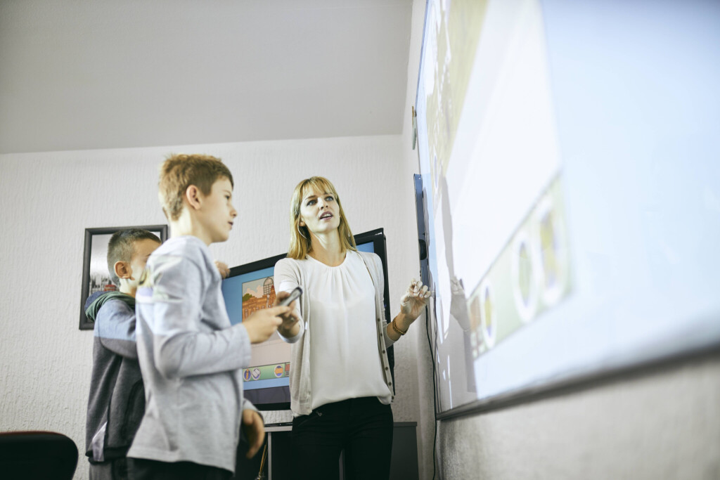 Eine Lehrerin reicht einem Schüler, der vor einem Whiteboard steht, einen Marker.