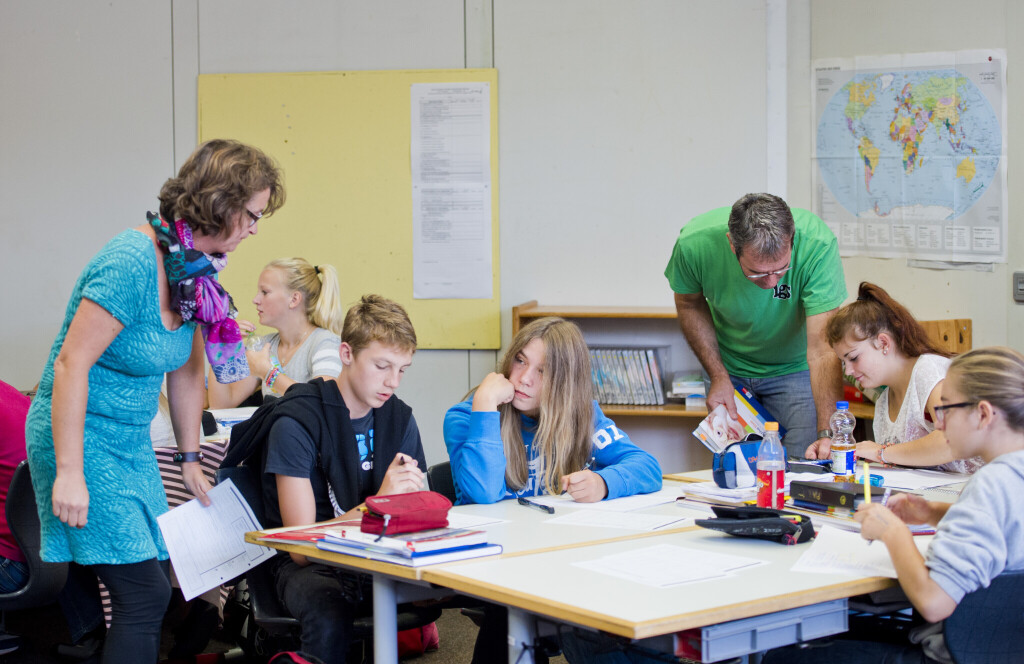 Ein Lehrer und eine Lehrerin stehen neben einer Tischgruppe und helfen den Schülern, die an den Tischen verteilt sitzen, bei ihren Aufgaben.