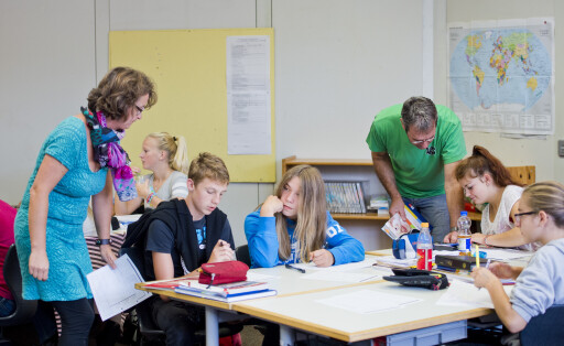 Ein Lehrer und eine Lehrerin stehen neben einer Tischgruppe und helfen den Schülern, die an den Tischen verteilt sitzen, bei ihren Aufgaben.