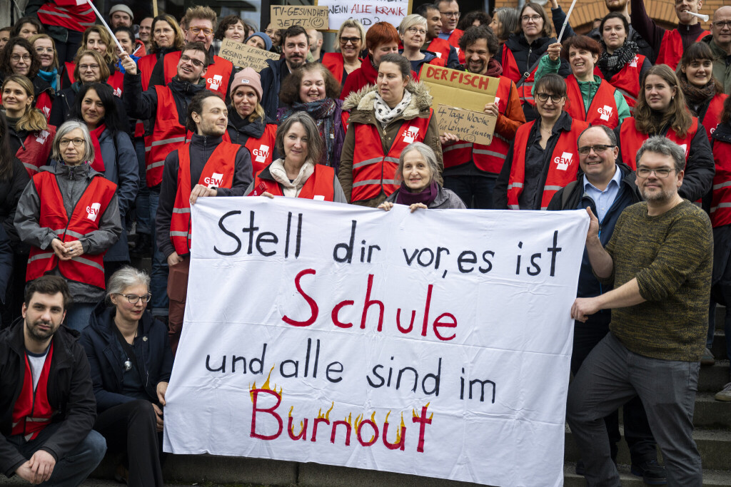In rote Warnwesten gekleidete Menschen stehen auf einer Treppe und halten ein selbstgemaltes Banner in der Luft, auf dem geschrieben steht: "Stell dir vor, es ist Schule und alle sind im Burnout".