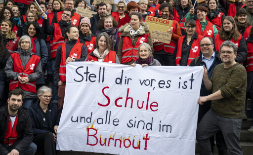 In rote Warnwesten gekleidete Menschen stehen auf einer Treppe und halten ein selbstgemaltes Banner in der Luft, auf dem geschrieben steht: "Stell dir vor, es ist Schule und alle sind im Burnout".