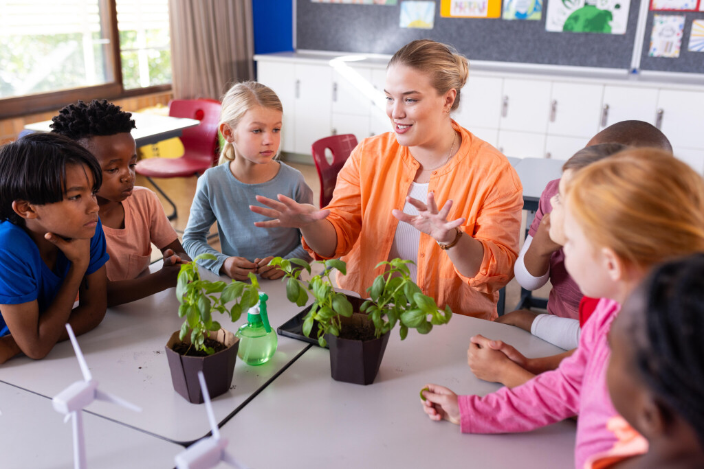 Eine junge Lehrerin sitzt mit sieben Schülern und Schülerinnen an einem Tisch, auf dem Pflanzen stehen.