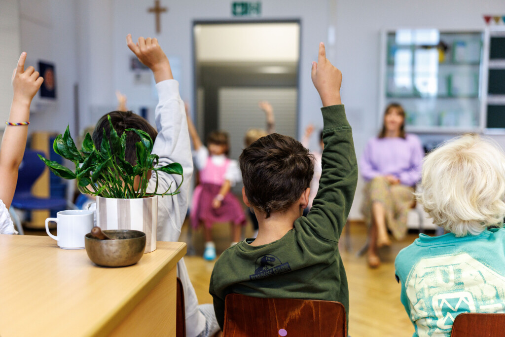 Grundschüler, die in einem Stuhlkreis sitzen, melden sich. Im Hintergrund sitzt eine Lehrerin.