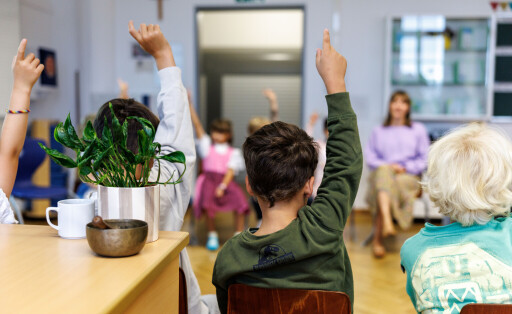 Grundschüler, die in einem Stuhlkreis sitzen, melden sich. Im Hintergrund sitzt eine Lehrerin.