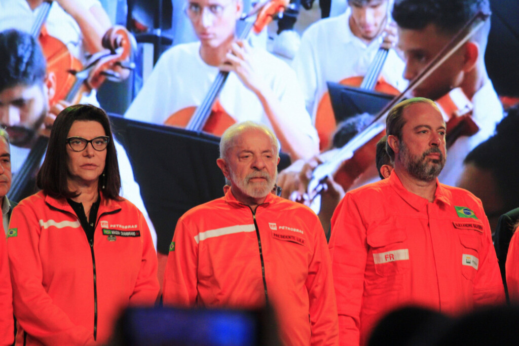 Drei Menschen stehen in leuchtend orangeroten Arbeitsanzügen auf einer Bühne, im Hintergrund spielt ein Streichorchester.