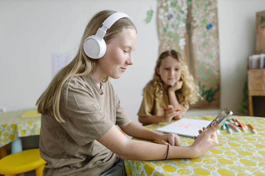 Girl taking a video call with headphones while sibling smiles nearby at a table in a cozy indoor setting